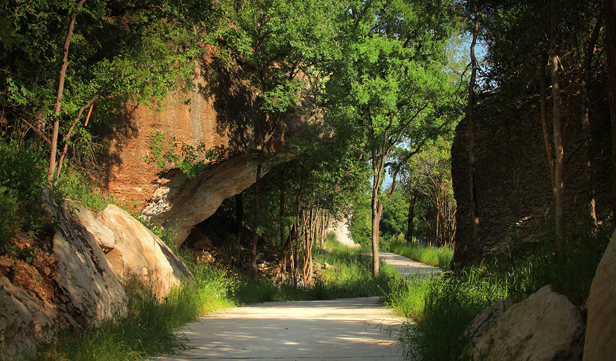 Hiking trail in the Wolf Ranch community, with lush green vegetation and a shaded pathway with vibrant orange dirt. Hiking trail in the Wolf Ranch community, with lush green vegetation and a shaded pathway with vibrant orange dirt.